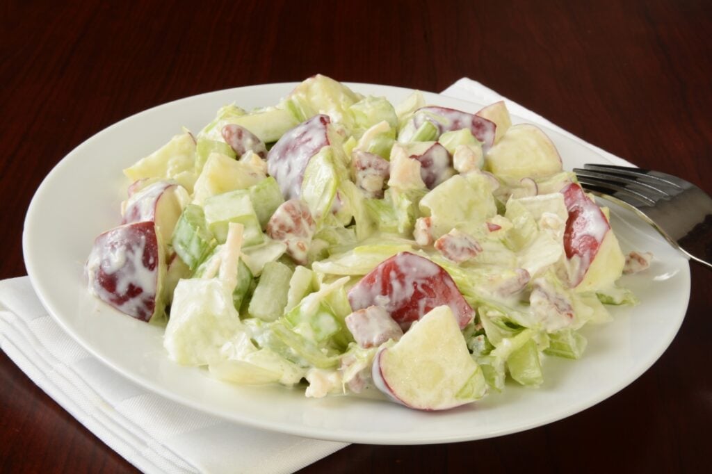 A plate of Waldorf salad with chopped apples, celery, walnuts, and lettuce in a creamy dressing, served on a white plate with a fork and napkin on a dark wooden table.
