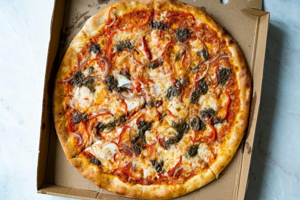 A whole pizza with cheese, tomato slices, red onion, and pesto, sitting in an open cardboard pizza box on a white surface.
