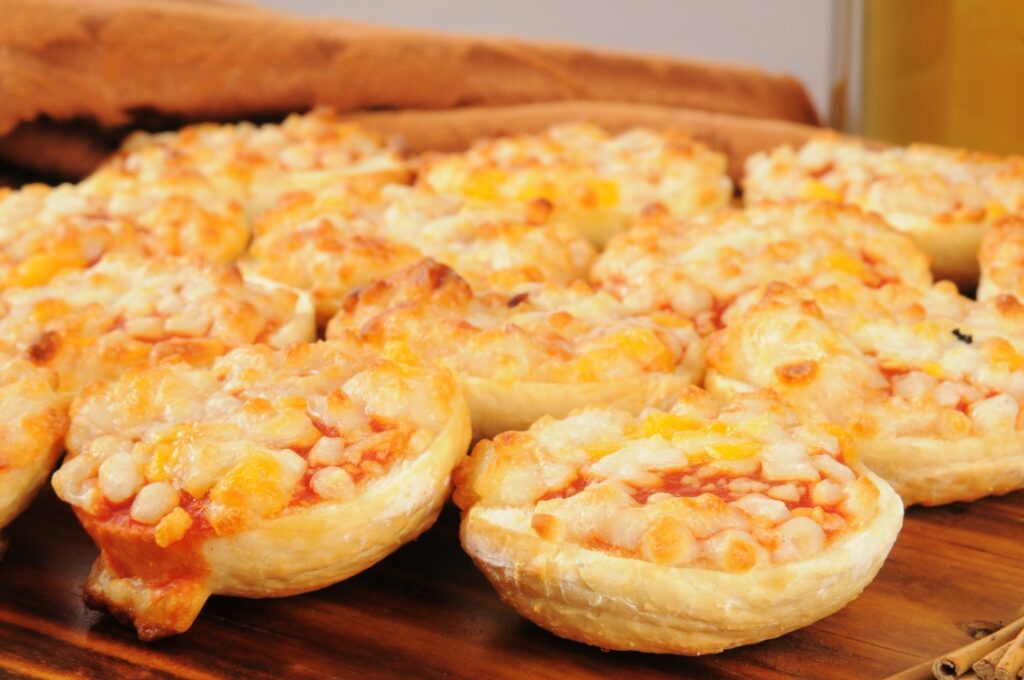 A close-up of several mini pizzas with melted cheese and diced toppings, arranged on a wooden surface with a brown cloth in the background.