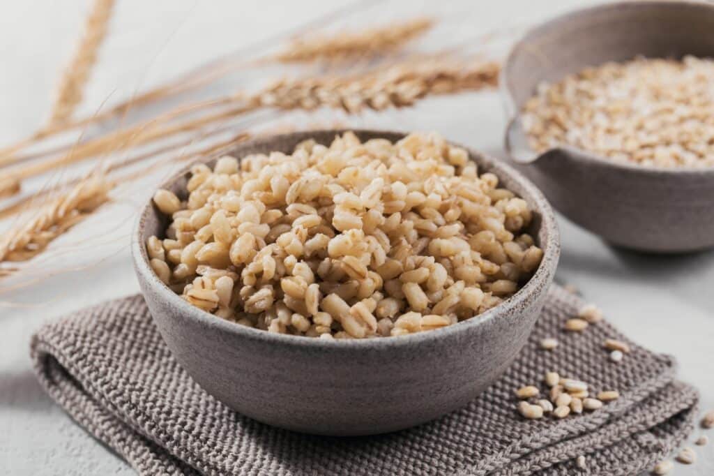 A gray bowl filled with cooked barley sits on a folded cloth, with wheat stalks and another bowl of uncooked barley in the background.