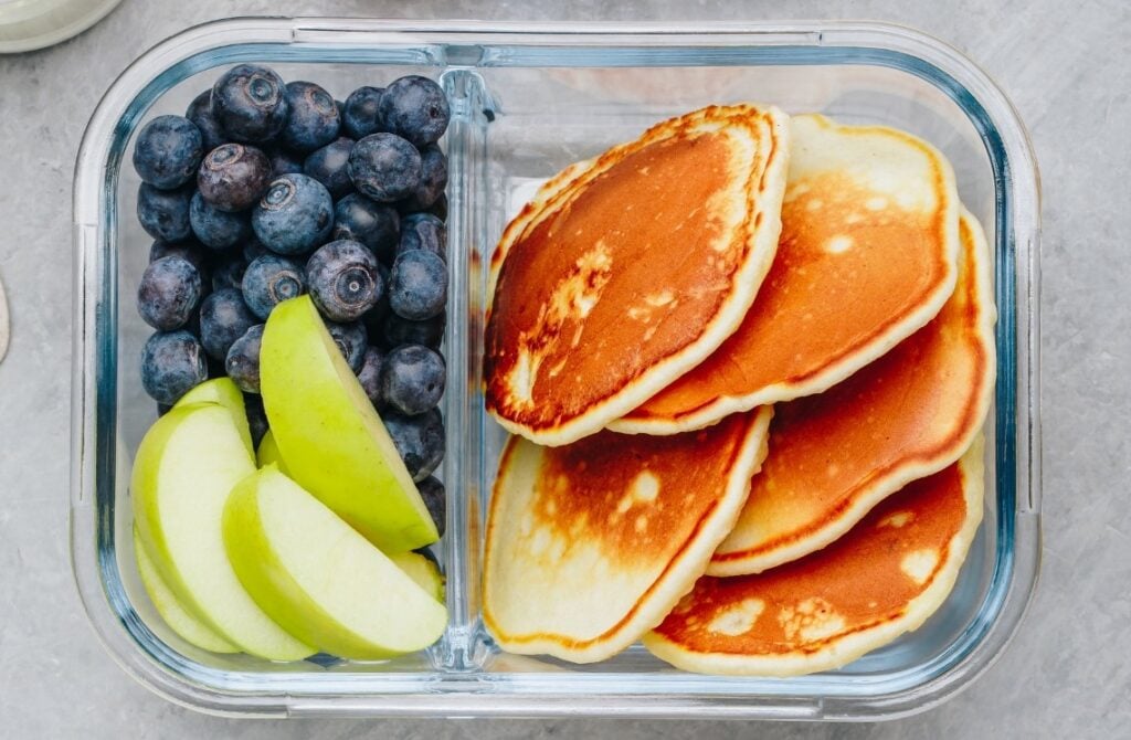 A glass container with three sections holding fresh blueberries, sliced green apples, and several golden-brown pancakes on a gray surface.