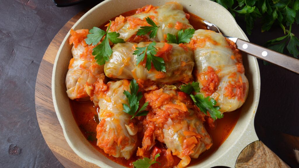 Cooked cabbage rolls in a tomato-based sauce, garnished with fresh parsley, served in a beige ceramic bowl with a fork.