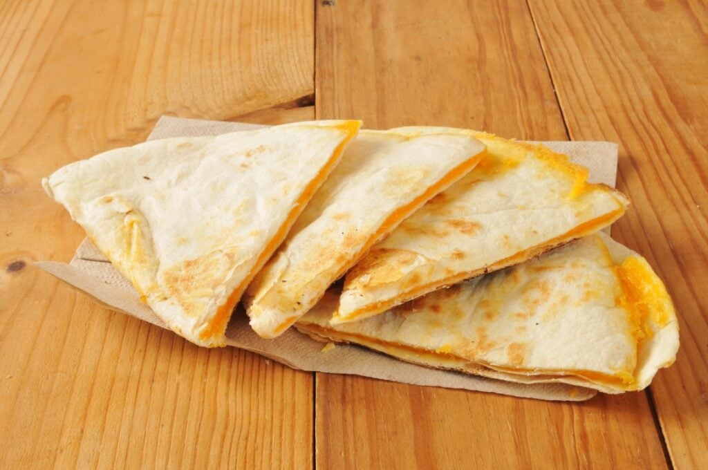 Four cheese quesadilla wedges with melted cheese are stacked on a brown napkin atop a wooden table.