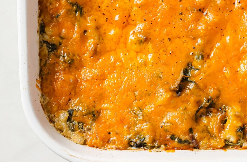 A close-up of a baked casserole in a white dish, topped with melted and slightly browned cheddar cheese, with bits of greens and other ingredients visible beneath the cheese layer.