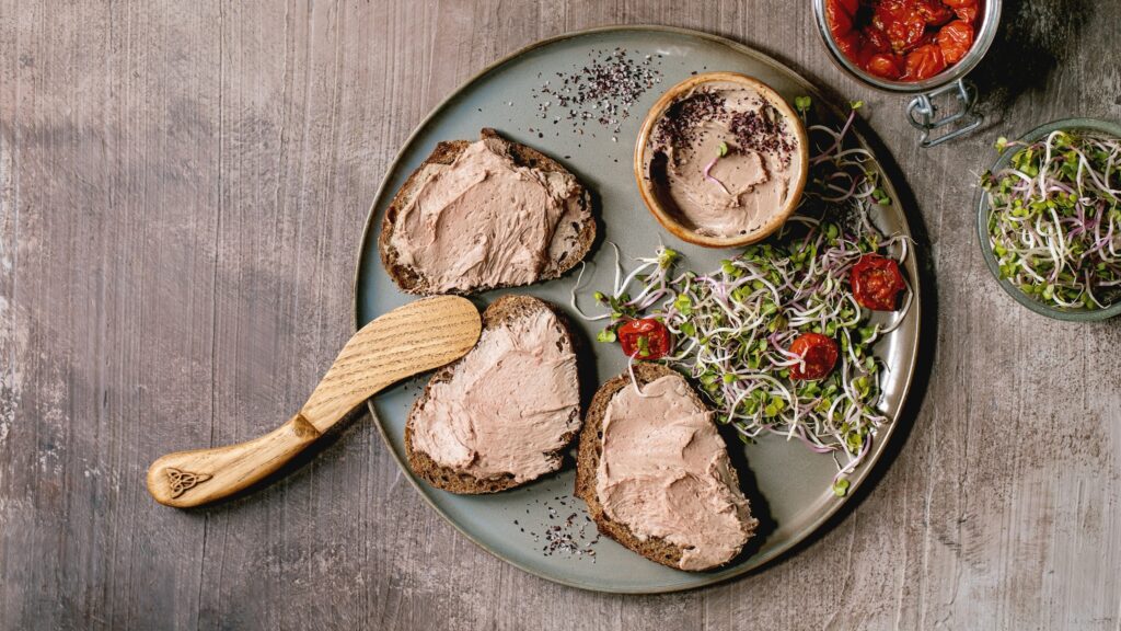 Three slices of rustic bread topped with chicken liver pâté on a plate with a wooden spreader, microgreens, and roasted tomatoes.