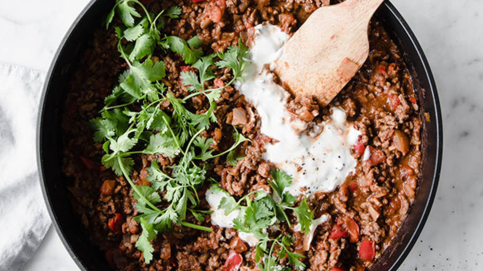 Skillet of chili con carne topped with sour cream and fresh cilantro, with a wooden spoon.