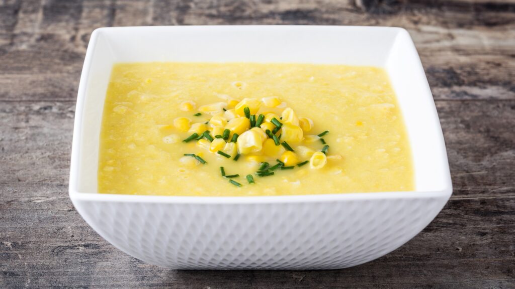 Smooth yellow creamed corn served in a white square bowl, topped with corn kernels and chopped chives on a wooden table.