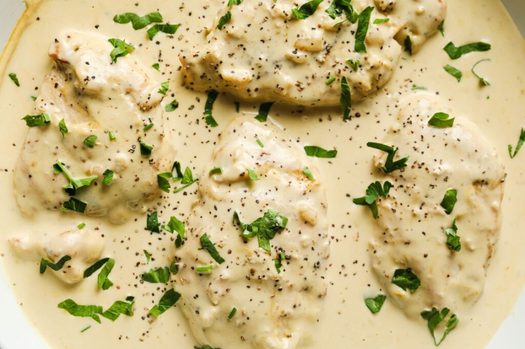 Four pieces of chicken breast covered in a creamy white sauce, garnished with chopped parsley and freshly ground black pepper.