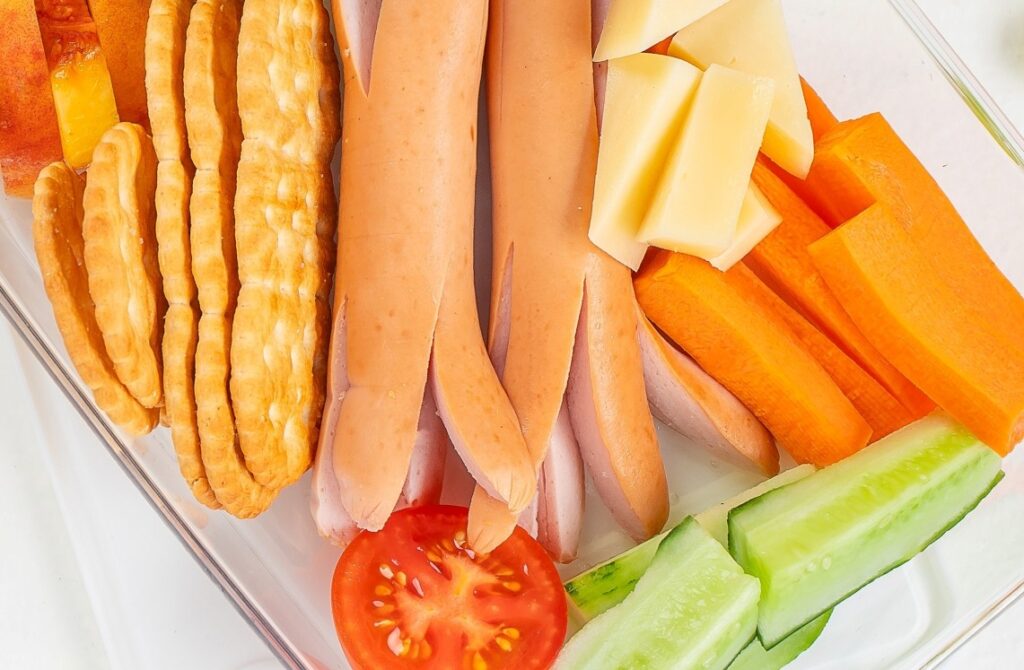 A plate with sliced sausages, cheese cubes, carrot sticks, cucumber slices, round crackers, a tomato slice, and a piece of peach arranged neatly.