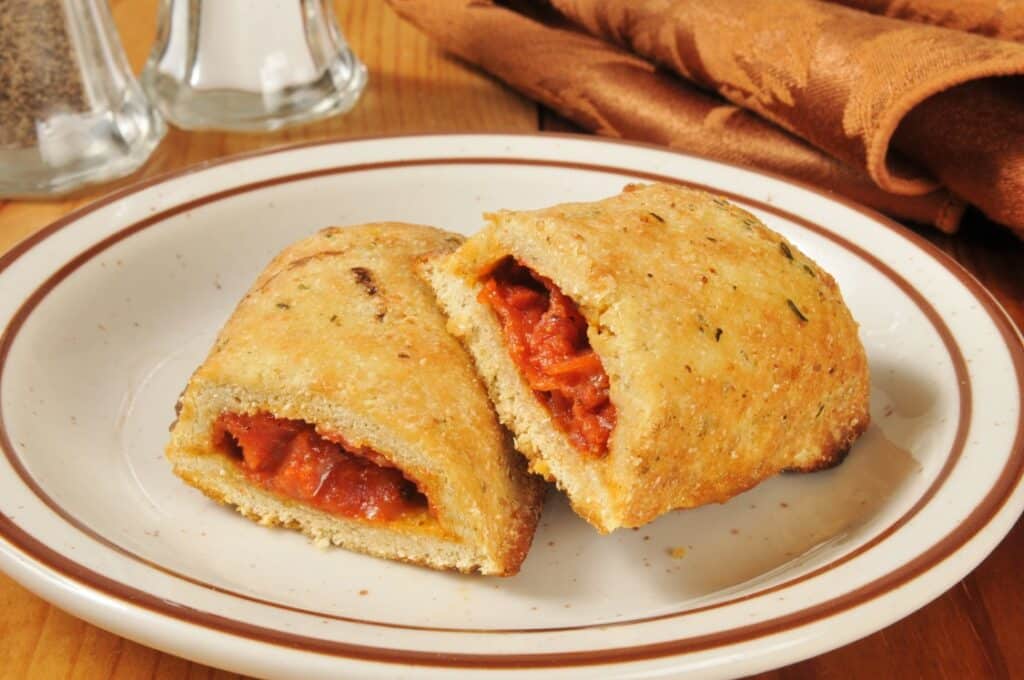 A plate with a baked calzone cut in half, showing a filling of tomato sauce, cheese, and pepperoni. A brown napkin and salt and pepper shakers are in the background.