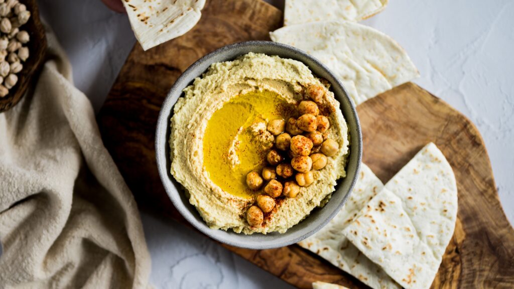 Creamy hummus served in a bowl, topped with whole chickpeas, paprika, and olive oil, surrounded by pieces of flatbread on a wooden board.