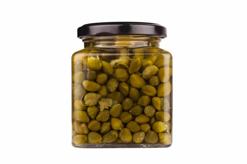A glass jar filled with green capers, sealed with a black lid, isolated on a white background.