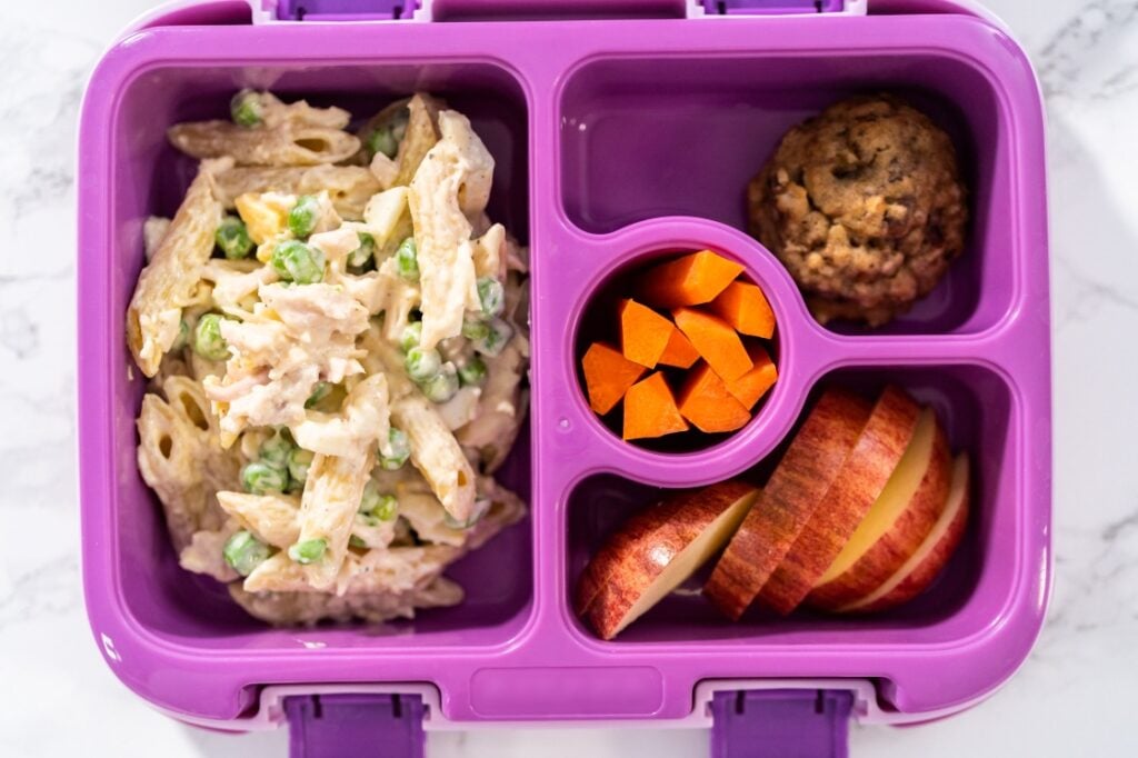 A purple lunchbox with four compartments holding creamy pasta with peas, carrot slices, a chocolate chip cookie, and apple slices.