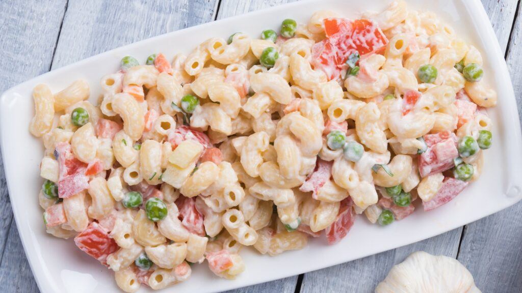 Macaroni salad with elbow pasta, peas, and diced vegetables in a creamy dressing, served on a white rectangular platter.