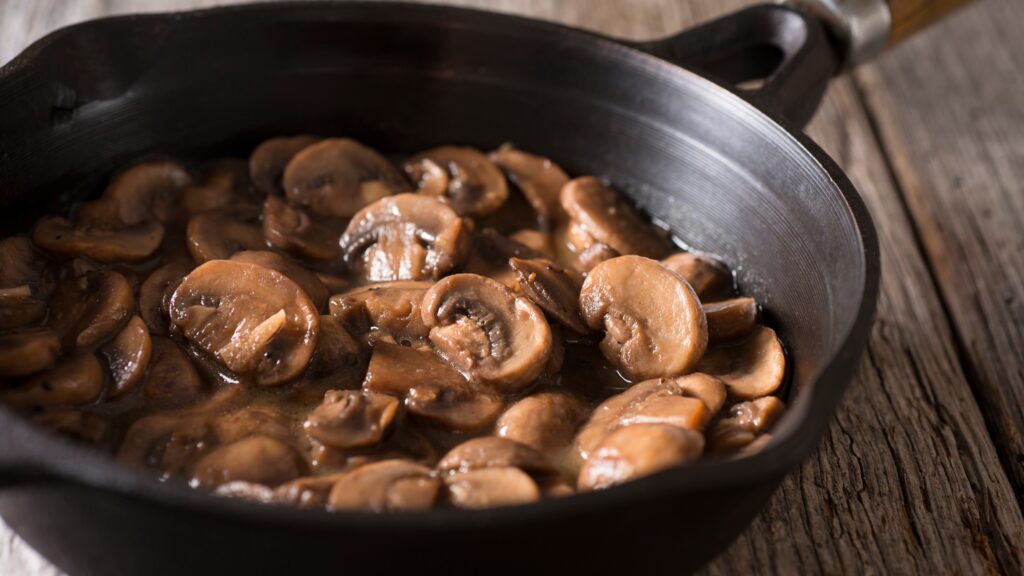 Sliced mushrooms sautéed in a skillet, glistening with a savory sauce, placed on a rustic wooden surface.