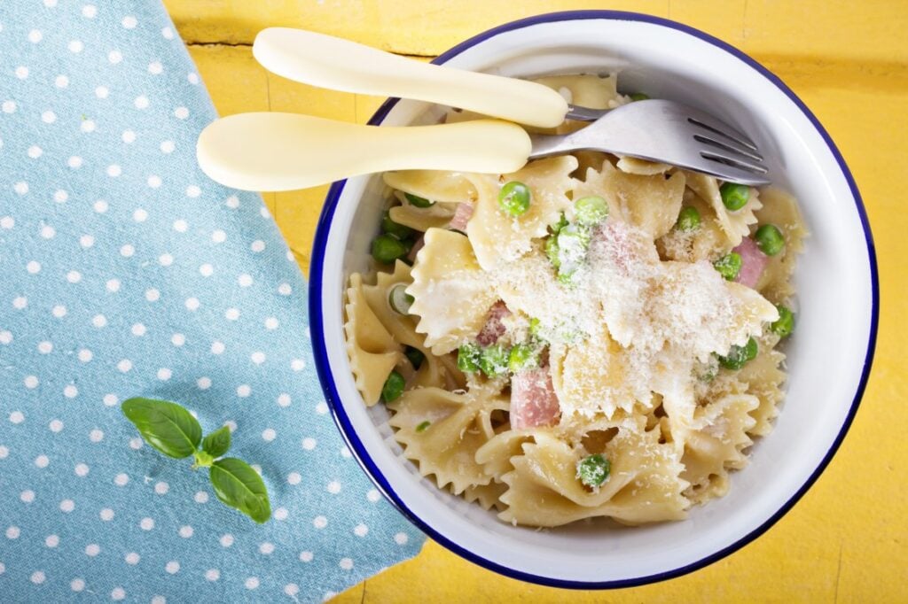 A bowl of farfalle pasta with peas, ham, and grated cheese, served with a fork and knife. The bowl is on a yellow surface next to a blue polka-dotted cloth and a sprig of basil.