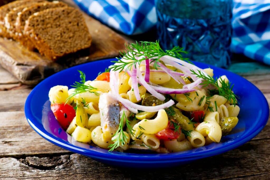 A blue plate with pasta salad topped with red onion slices, dill, tomatoes, and pieces of fish. Slices of whole grain bread are in the background on a wooden surface, with a blue glass and checkered cloth nearby.