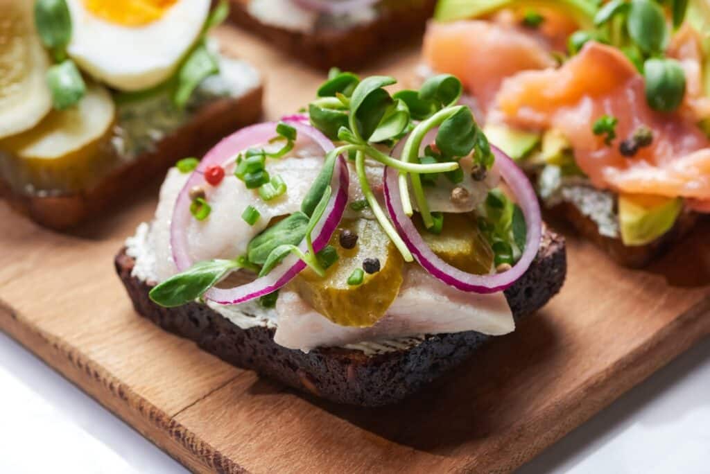 An open-faced sandwich on dark bread topped with pickled herring, red onion rings, pickles, microgreens, and peppercorns, on a wooden board with other assorted sandwiches in the background.