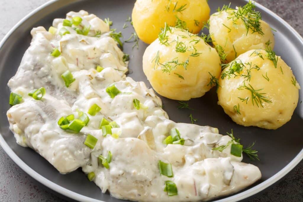 A gray plate with herring fillets in creamy white sauce garnished with chopped green onions, served alongside three boiled potatoes topped with fresh dill.