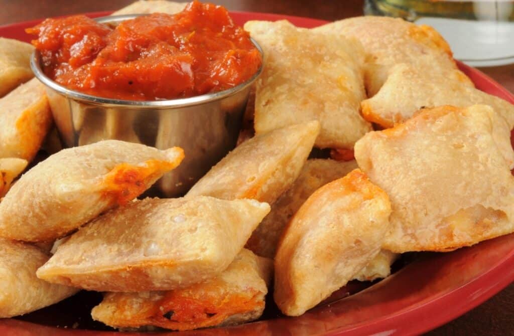 A plate of golden-brown pizza rolls surrounds a metal cup filled with marinara sauce for dipping. The rolls appear crispy, with hints of red tomato sauce and cheese visible inside.