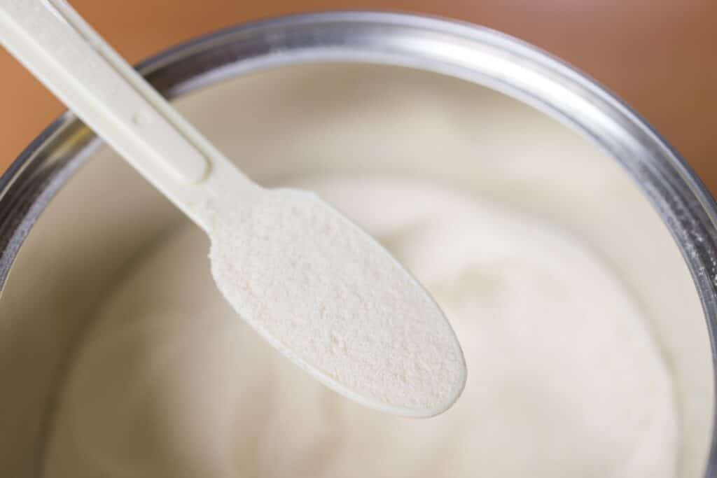 A close-up of a plastic scoop filled with powdered infant formula, held above an open container of the same formula.