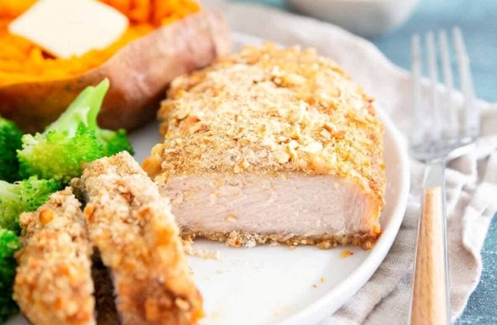 A plate with a piece of breaded baked chicken breast, slices of the chicken, steamed broccoli, and a baked sweet potato with a pat of butter. A fork rests on the plate next to the food.