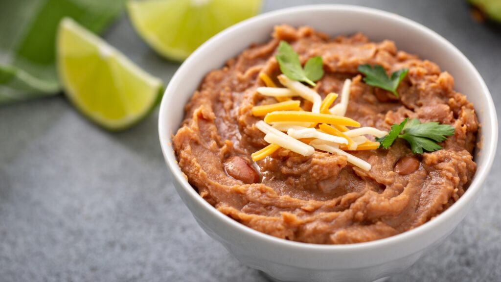 Bowl of refried beans topped with shredded cheese and cilantro, surrounded by lime wedges on a gray surface.