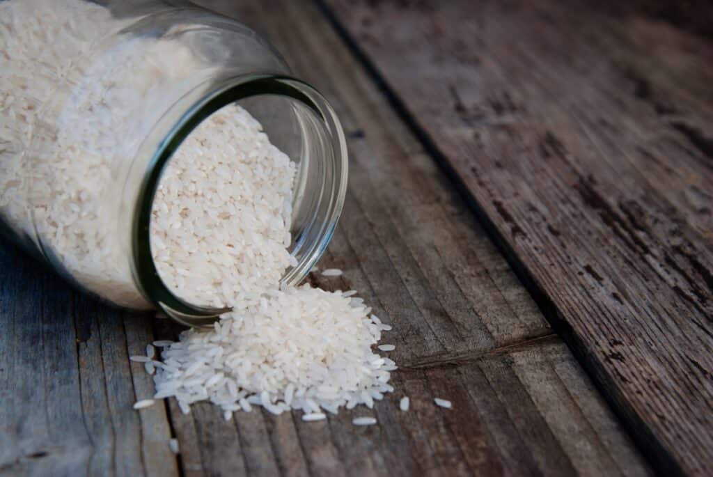 A glass jar tipped over on a rustic wooden surface, spilling white rice grains onto the table.