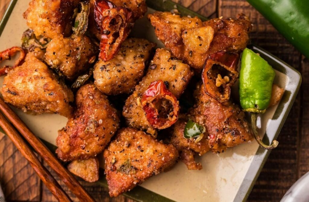 A plate of crispy pork chop pieces garnished with sliced red and green chili peppers, black pepper, and herbs, served on a square plate with wooden chopsticks nearby.