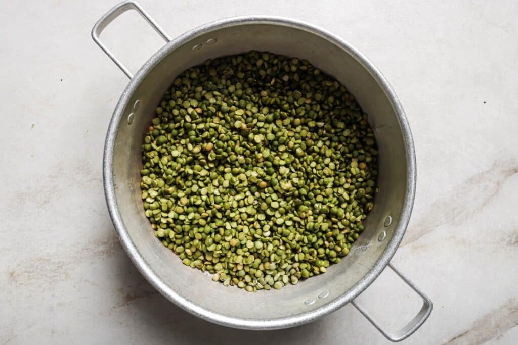 A metal pot with handles on both sides is filled with dry green split peas, placed on a light-colored countertop.