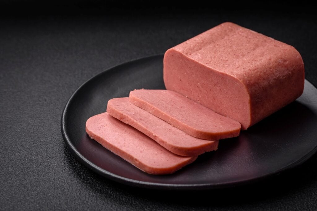A block of pink processed meat with three rectangular slices cut, arranged on a round black plate against a dark background.