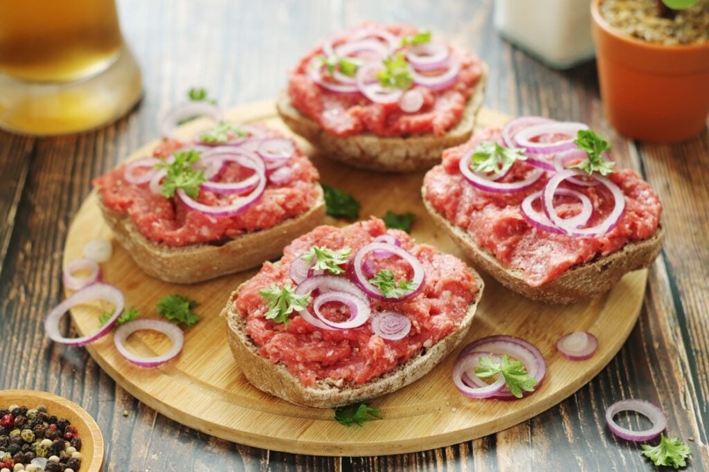 Four pieces of rye bread topped with raw minced meat, sliced red onions, and parsley are arranged on a round wooden board on a rustic table. Spices, a drink, and potted herbs are in the background.