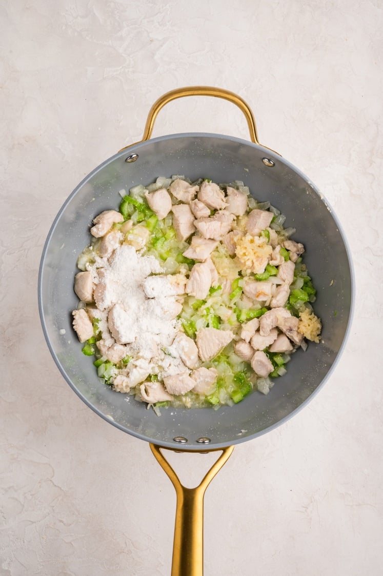 A pot with chopped chicken, diced celery, onions, minced garlic, and a sprinkle of flour, all being sautéed on a light-colored surface.