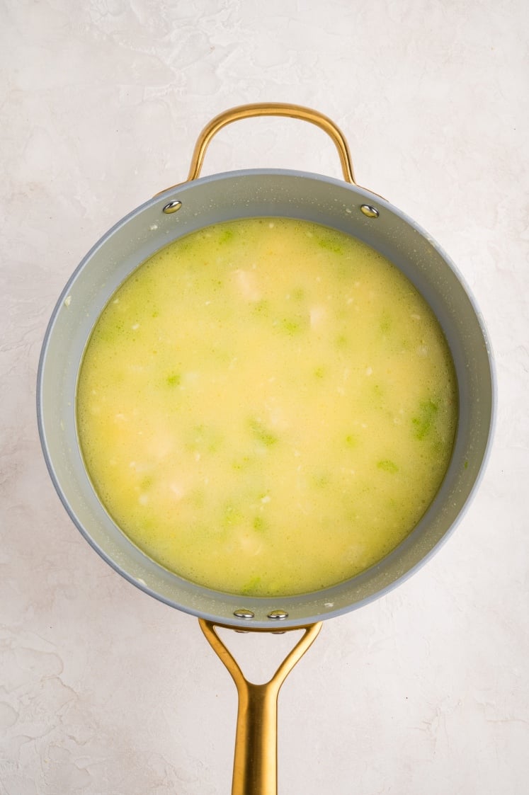 A pot of light yellow soup with small green and white pieces, seen from above, sits on a light, textured surface. The soup appears to be simmering gently.