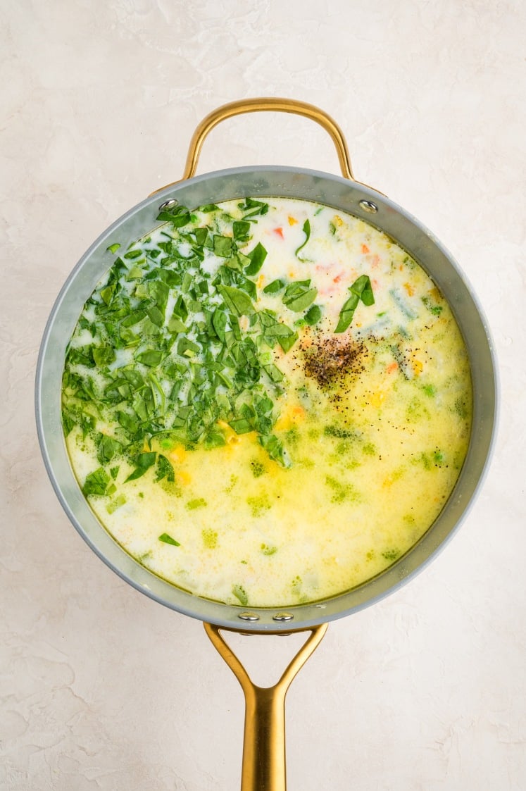 A pot of creamy soup on a white surface, containing chopped spinach, visible black pepper, herbs, and a light yellow broth, with a golden handle on the pot.