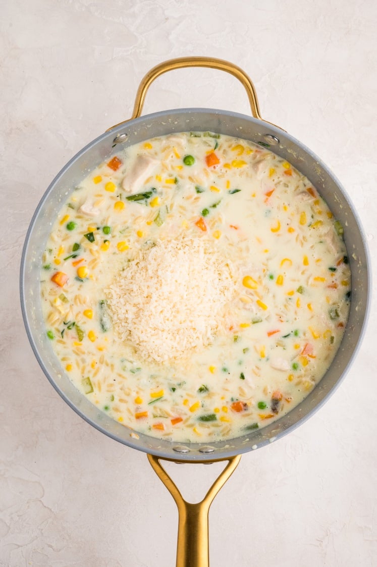 A large saucepan filled with creamy orzo pasta containing corn, carrots, green beans, and chicken, topped with a mound of shredded cheese ready to be stirred in.