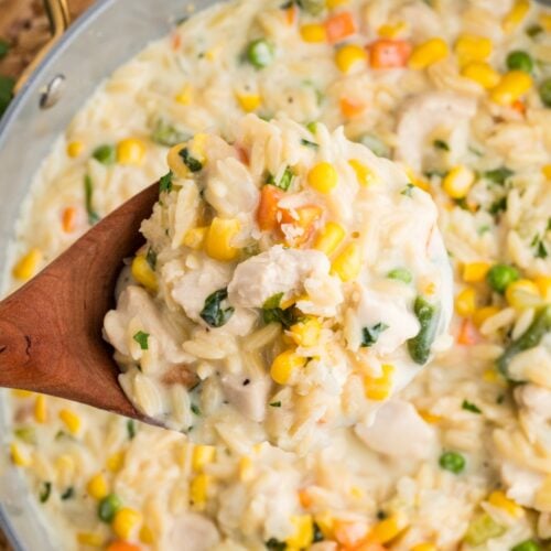 A wooden spoon holds a serving of creamy orzo pasta mixed with peas, corn, carrots, and small pieces of chicken over a large pan filled with the same mixture.