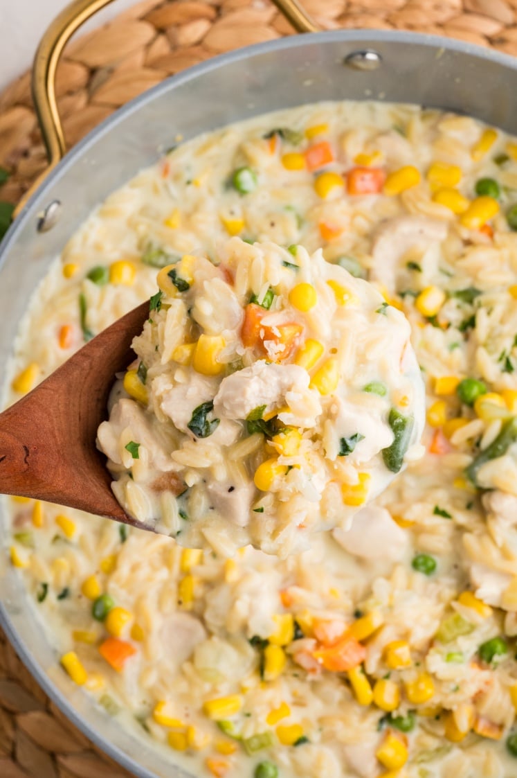 A wooden spoon holds a serving of creamy orzo pasta mixed with peas, corn, carrots, and small pieces of chicken over a large pan filled with the same mixture.
