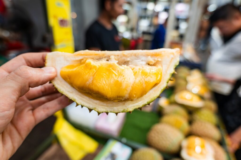 A hand holds a freshly cut piece of ripe durian fruit at an outdoor market stall, with whole durians and blurred people visible in the background.