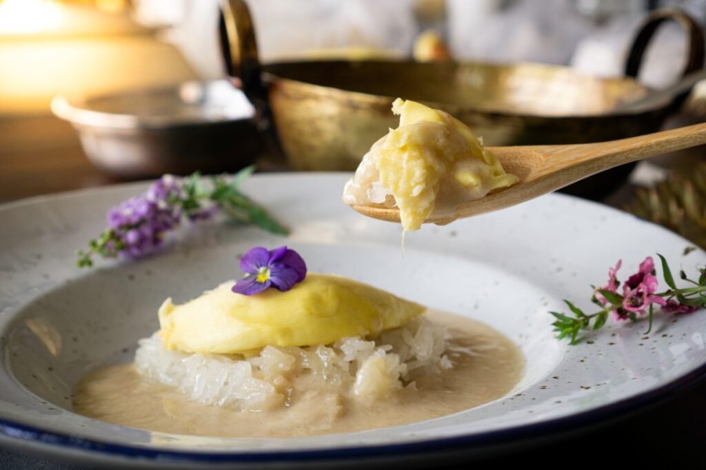 A plate of sticky rice topped with durian and coconut sauce, garnished with a purple edible flower, is shown with a wooden spoon lifting a portion. Decorative flowers are placed beside the dish.