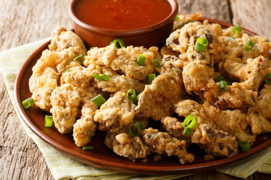 A plate of crispy fried oyster mushrooms garnished with chopped green onions, served with a small bowl of red dipping sauce on a wooden surface.