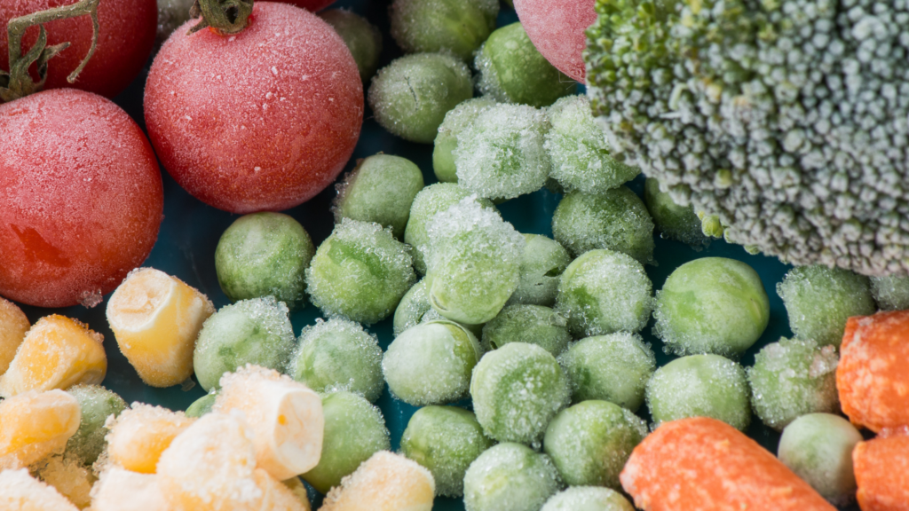 Close-up of assorted frozen vegetables including tomatoes, peas, carrots, corn, and broccoli, covered in frost.