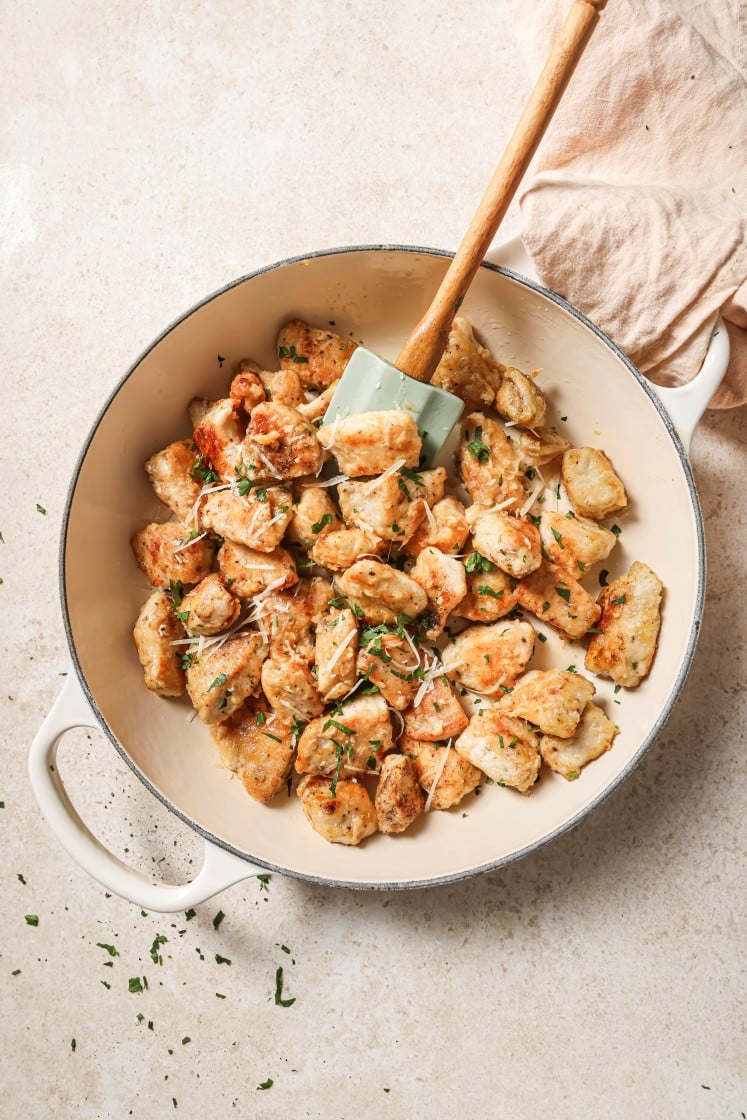 A white Dutch oven filled with golden, pan-seared gnocchi, garnished with chopped herbs and grated cheese. A spatula rests in the pot, with a beige cloth nearby on a light countertop.