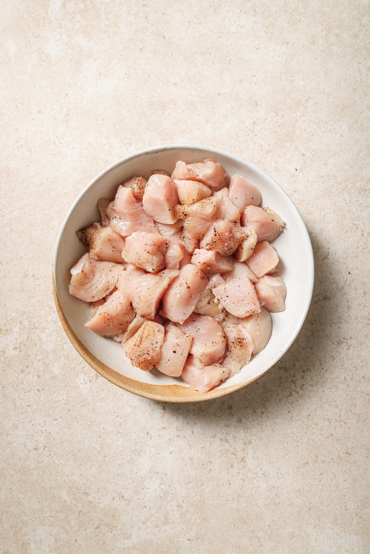 A white bowl filled with raw, diced chicken pieces seasoned with black pepper, placed on a light beige surface.