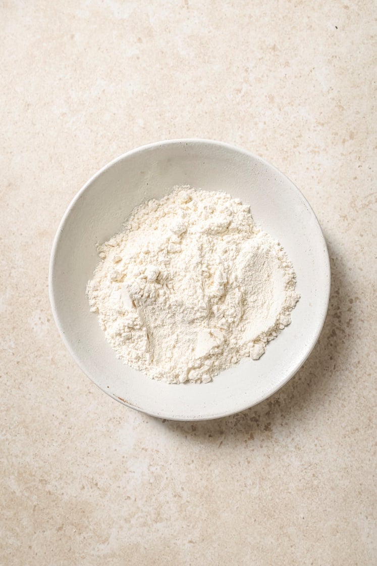 A white ceramic bowl filled with a mound of white flour sits on a light beige textured surface.