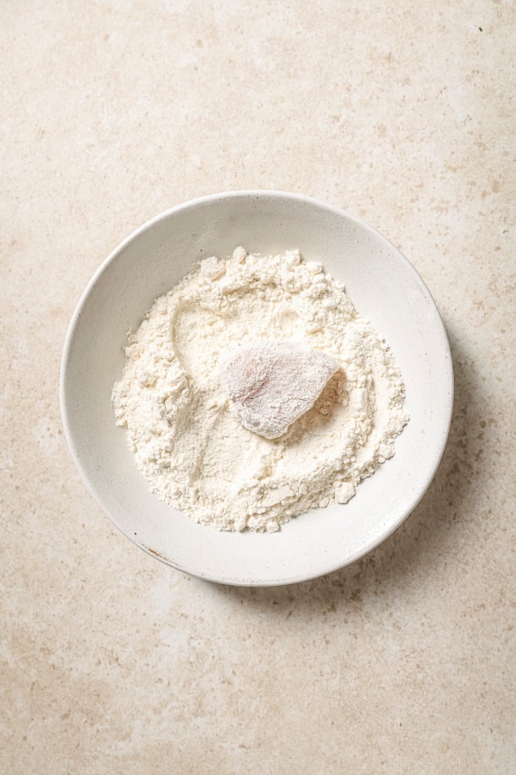 A bowl filled with flour and a piece of raw meat partially coated in the flour, placed on a light-colored countertop.