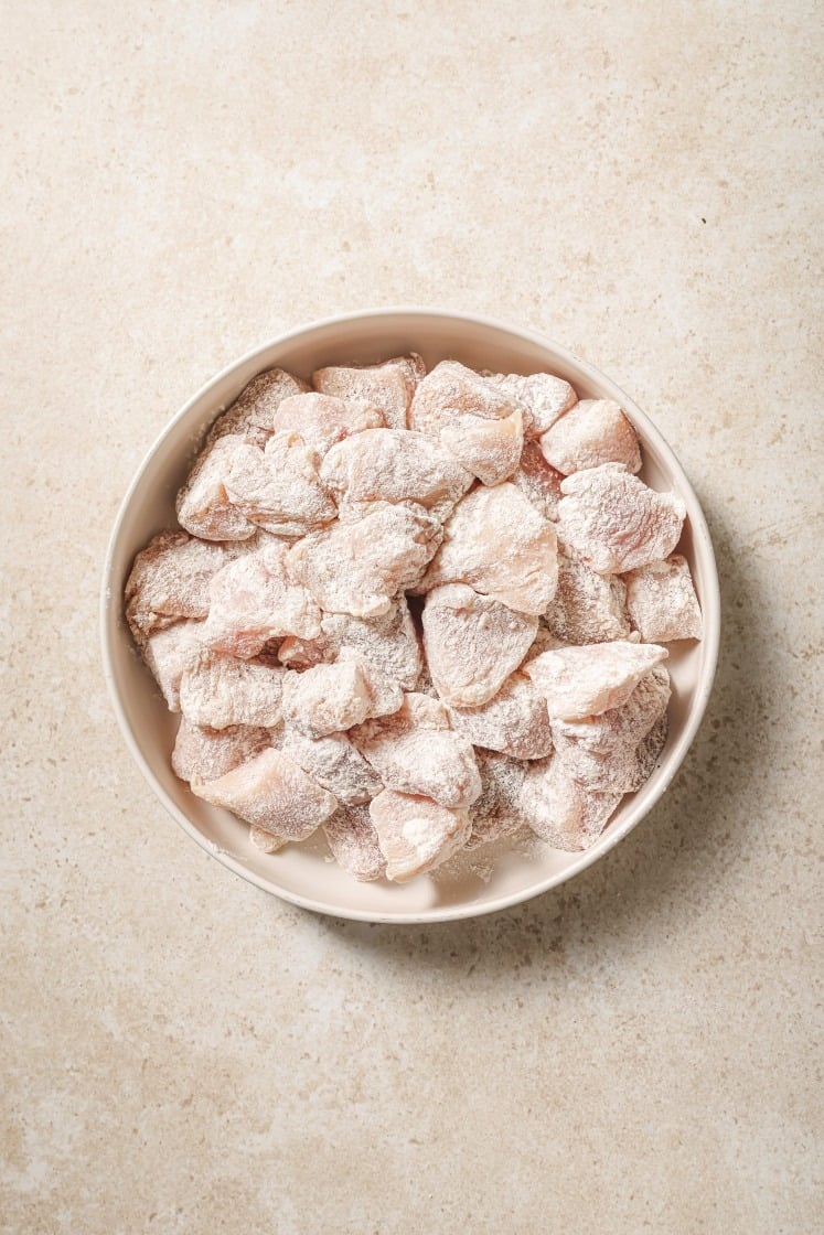 A bowl filled with raw chicken pieces coated in flour or starch, placed on a light, textured surface.