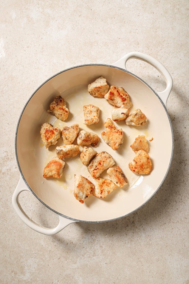 Chunks of cooked chicken breast are spread out in a white, round, enameled pot with two handles, sitting on a light-colored countertop.