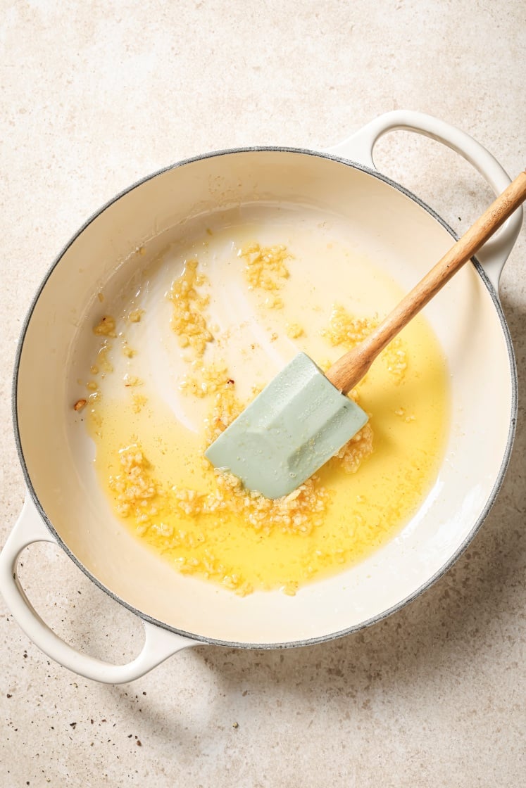 A white pot with sautéed minced garlic in melted butter sits on a light countertop. A light green silicone spatula with a wooden handle rests inside the pot.