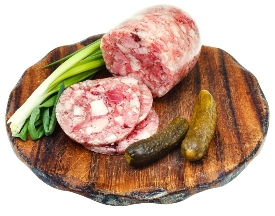 A wooden board with a round loaf of head cheese, two slices of it, two whole pickles, and several green onions, all arranged neatly.